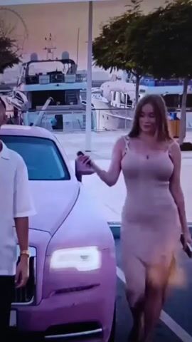 A man and a woman are standing next to a pink Rolls-Royce Phantom. The man is wearing a white button-down shirt and black pants. The woman is wearing a beige dress with a slit up the side. The car is parked in front of a body of water with several boats and yachts docked. In the background, there is a Ferris wheel. The sky is a soft pink and orange, indicating sunset or sunrise. The man appears to be looking at the woman, who is looking forward.
