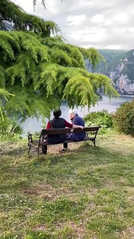 The video captures two older individuals seated on a dark wooden park bench, positioned on a grassy hill overlooking a stunning natural landscape. A large tree with vibrant green foliage partially frames the left side of the shot, its branches extending towards the bench. The person on the left, seen from behind, wears a dark vest over a red long-sleeved shirt. The person on the right, with white hair, wears a dark blue jacket, and the person on the left gently places a hand on their arm.

Beyond the bench, a serene body of water, likely a lake or river, winds through a dramatic canyon. Towering mountains, covered in lush green vegetation and with visible rocky outcrops, rise majestically on both sides of the water. The sky is partly cloudy, with soft, diffused light suggesting late afternoon or early evening. The foreground is a well-maintained grassy area. The scene conveys a sense of peace and companionship amidst breathtaking natural beauty. There is no visible text. The location context indicates this is in Plužine, Montenegro, renowned for its dramatic landscapes like the Piva Lake and canyon.