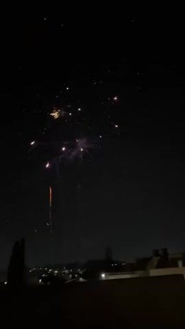 This is a video of fireworks being set off at night in Vila Nova de Gaia, Portugal. The fireworks are a variety of colors including gold, white, and pink, and they explode in the sky creating bright, starburst patterns. Some of the fireworks leave trails of smoke as they fall. In the foreground, the silhouettes of people can be seen watching the display. The bottom of the frame shows the rooftops of buildings in Vila Nova de Gaia, with some lights visible in the distance, suggesting an urban or suburban setting. The sky is dark, indicating it is nighttime. The overall mood of the video is celebratory and exciting, due to the vibrant fireworks display. There is no visible text in the video.