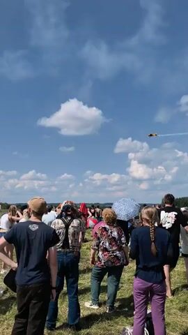 This is a vertical, low-angle shot from a ground-level perspective at what appears to be an outdoor event or airshow. A crowd of people, viewed from behind, are looking up at the sky. The sky is bright blue with scattered white clouds, and in the distance, two yellow-colored propeller planes are performing aerial maneuvers, leaving white smoke trails in their wake. The people in the foreground are casually dressed for warm weather, and some are holding umbrellas or wearing hats. The ground is covered in dry grass, with a few scattered items like chairs and tents visible. The overall atmosphere suggests a sunny day and an event focused on aviation or entertainment.