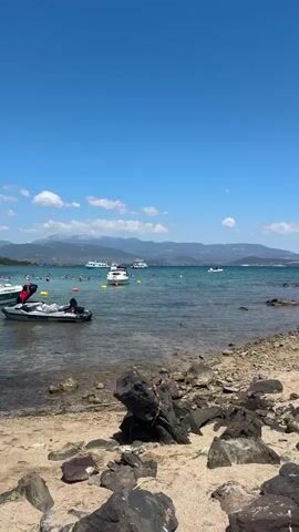 The video shows a coastal scene with a clear blue sky. In the foreground, there is a rocky beach with some driftwood. The water is turquoise and calm, with several boats and jet skis visible. In the distance, there are mountains. The overall atmosphere is peaceful and idyllic.