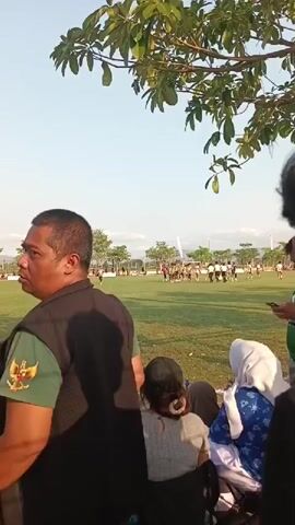A group of people are gathered on the side of a grassy field, watching a soccer game in progress. In the foreground, a man in a dark vest with an emblem on the sleeve stands to the left. To the right, a person with dark hair is partially visible, facing away from the camera. In the middle ground, two people are seated, facing the field. The soccer game is visible in the distance, with players on the field and spectators along the sidelines. The environment appears to be outdoors, with trees and clear sky visible. The overall atmosphere suggests a sporting event.