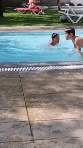 A group of children are playing in a swimming pool. One child is partially submerged in the water, and another child is on their back in the pool. The child on top is looking at the camera, and the child underneath is submerged with only their head visible. In the background, there are lounge chairs and people relaxing by the pool. The pool is surrounded by a tiled patio.