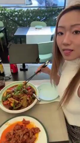 A woman with long blonde hair sits at a restaurant table enjoying a meal. In front of her are two plates of food: one large bowl containing a mix of meats and vegetables in a dark sauce, and a smaller plate filled with what appears to be pieces of meat in a reddish sauce. She uses chopsticks to pick up a piece of food and eat it. A tablet stands in front of her on a black stand. In the background, a window offers a view to the outside street, and a restaurant sign is visible.