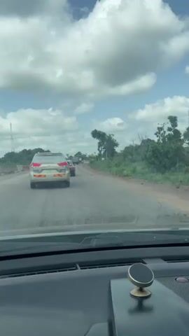 The video depicts a view from inside a car as it travels along a paved road. The time of day appears to be daytime, with bright sunlight and scattered clouds in a blue sky. The weather seems pleasant, with fluffy white clouds suggesting fair conditions. The road is flanked by lush green vegetation and trees, indicating a rural or semi-rural setting.

In the foreground, the dashboard of the car is visible, including a car mount for a phone with a gold-colored magnetic holder. The primary subject in the video is a silver SUV driving ahead in the same lane. The SUV has a license plate and stickers on its rear window. One sticker, visible on the right side of the rear window, appears to be text-heavy. The car ahead also has a visible antenna and a sunroof.

Further down the road, other vehicles are faintly visible, including a red car and what might be a truck. The road itself shows signs of wear, with some cracks and uneven patches. To the left of the road, there are concrete barriers, suggesting ongoing construction or road work. In the distance, a tall telecommunications tower is visible against the sky.

The overall mood is one of travel and movement, with a sense of journeying through a natural landscape. The presence of construction barriers might imply that the road is undergoing improvements. The location context provided, Aciol, Nigeria, places the scene in a specific geographic region, contributing to the understanding of the environment depicted.