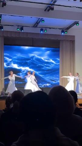 The video shows five young female ballet dancers on a stage in front of an audience. They are all wearing long white dresses and are performing a dance with their arms outstretched. Behind them is a large screen showing a digital image of crashing ocean waves. The stage is lit with blue lights. The audience is visible in the foreground, mostly consisting of people with grey or white hair.