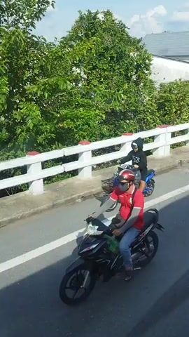 The video shows a view from a moving vehicle, likely a bus or car, traveling along a road in An Hữu, Vietnam. The road is paved with white lane markings. On the right side of the road, there's a raised concrete barrier with white railings and red accents, separating the road from a vegetated area with trees and some buildings.

Several motorcycles are visible. In the foreground, a person wearing a red shirt, blue jeans, a red helmet, and a black face mask rides a black motorcycle. They are wearing grey arm sleeves. Further down the road, another motorcycle is visible, and a third motorcycle is seen in the distance.

The scene appears to be daytime, with bright sunlight casting shadows. The weather seems clear and sunny, indicated by the bright light and blue sky with some clouds visible through the trees.

Along the roadside, there are various signs and structures. Some signs have Vietnamese text. One sign appears to be for a business, with "HƯƠNG VIỆT" visible. Another sign has "ONG MAP" written on it. There are also displays of goods, possibly for sale, such as what looks like coconuts or dried palm leaves lined up along the road. Banana trees are also visible in the background.

The overall impression is of a typical day of travel along a road in a rural or semi-rural area of Vietnam, with local transportation and commerce in evidence.