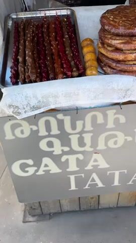 The video shows a close-up of a street vendor's stall, featuring various Armenian sweets and jams. On the left, several stacks of large, round, flatbreads or cakes with decorative patterns are visible. In the center, there's a sign that says "GATA" in both Armenian and English, referring to the traditional Armenian sweet bread. Behind the sign, a variety of colorful churchkhela (nut-and-fruit candy) hangs in long strings. To the right and behind the churchkhela, numerous jars of homemade jams and preserves are stacked. The stall's counter is covered with a white lace tablecloth. Throughout the video, a person's hands can be seen briefly, indicating a transaction or interaction with the goods. The overall setting appears to be an outdoor market or street stall.