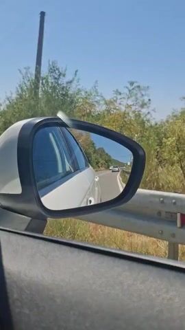 The video shows a view from the passenger side of a car. The camera is pointed towards the side mirror, which reflects the road behind the car and the surrounding environment. The car is driving on a paved road that curves to the right. There are trees and foliage on both sides of the road. A guardrail is visible alongside the road. In the distance, another car can be seen on the road. The sky is clear and blue, suggesting a sunny day. The overall scene depicts a scenic drive.