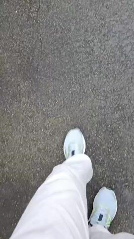 A first-person perspective video shows someone walking on a paved path through a lush, green environment. The path is dark and appears wet in some areas, possibly from rain. On either side of the path are dense trees and foliage, with some areas showing bamboo-like plants. The path curves slightly as it leads into the distance, where more trees and vegetation can be seen, with sunlight filtering through the leaves. The person is wearing white pants and light blue athletic shoes with green accents. The video begins with a close-up of the person's feet on the ground before panning up to show the path and the surrounding greenery.