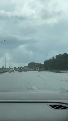 The video is taken from inside a car driving on a highway. The sky is overcast with dark clouds. The road is wet, suggesting it is raining or has recently rained. Several cars and trucks are visible on the highway in front of the car. The windshield wipers are visible in the foreground. Trees line the side of the highway.