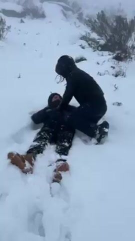 The video shows two people in a snowy environment. One person is lying on their back in the snow, while the other person kneels over them, appearing to interact with or assist them. The person on the ground is wearing a black jacket, patterned pants, and tan-colored boots. The other person is also wearing a black jacket with the hood up, dark pants, and black boots. The background consists of a snow-covered landscape with rocks and some sparse vegetation. It is snowing, and the visibility is somewhat limited due to the weather conditions. The person lying down struggles a bit to get up.