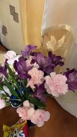 A close-up shot of a floral arrangement featuring pink carnations and purple alstroemeria lilies is presented. The flowers are in a blue vase, partially visible at the bottom left. The background consists of a beige patterned curtain or fabric. A portion of a yellow and red wrapper, possibly of a snack item, is visible in the lower left corner of the frame. The lighting is soft, suggesting an indoor setting. The video is composed of static frames, with no discernible movement or action occurring.