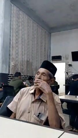 The video shows an elderly man sitting at a table in what appears to be a public space, possibly a cafe or restaurant. He is wearing a beige shirt, a black cap, and a ring on his finger. He is holding a lit cigarette and looking up, seemingly lost in thought. In the background, several other people are seated at tables, with some of them appearing to be engaged in conversation or using their phones. The room has striped wallpaper on one wall, and there is a small television and other electrical fixtures on another wall. A bicycle is also visible leaning against the wall in the background. The atmosphere seems casual and relaxed.