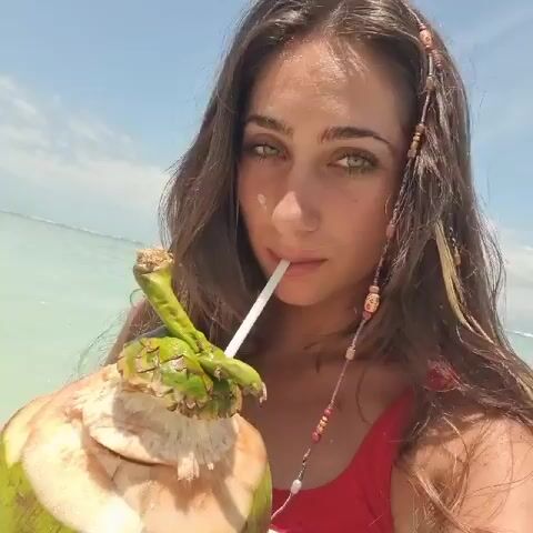 A woman in a red swimsuit stands in shallow, light green ocean water, holding a green coconut with a straw in it. She is looking at the camera and taking a sip from the coconut. She has long, brown hair with beaded strands and is smiling slightly. The sky is blue with scattered clouds.