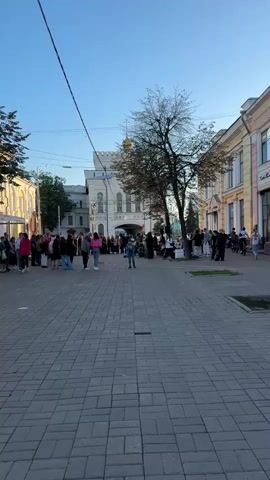 The video shows a street scene with a group of people gathered in front of a building. A young person in a blue jacket and jeans is standing in the foreground, seemingly dancing. Behind them, a band is performing, with several musicians playing instruments. Other people are watching the performance or walking by. The environment appears to be an urban street with a paved walkway, trees, and buildings in the background, one of which has a golden dome. The lighting suggests it is daytime, likely late afternoon or early evening.