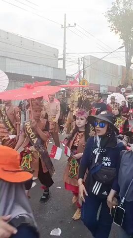 A group of people are participating in a parade. Many are dressed in traditional costumes with elaborate headwear and colorful fabrics. Some are carrying red and white umbrellas, while others hold banners and flags. There is a person on a scooter in the foreground. The parade appears to be taking place on a street with buildings lining the sides. The sky is overcast.