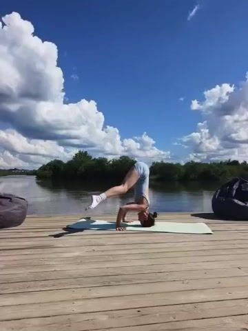 A woman is practicing yoga on a wooden deck by a body of water. She is in a headstand pose, with her legs extended upwards. The sky is blue with fluffy white clouds, and there are trees and foliage in the background. There are also two beanbag chairs visible on the deck.