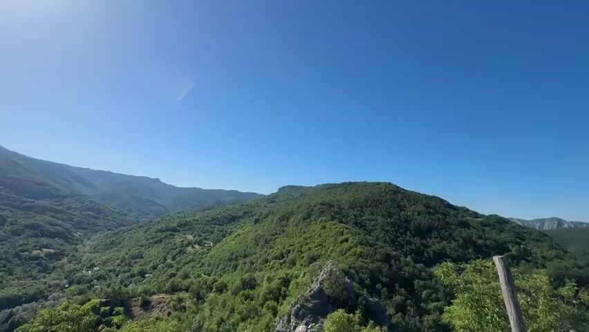 A wide shot of a lush green mountain landscape under a clear blue sky. The camera pans to reveal a wooden railing in the foreground, with two young women standing behind it, looking out at the view. One woman, with long dark hair, is holding up a selfie stick with a phone to take a picture. The other woman, with blonde hair pulled back in a ponytail, is looking down at her phone. The scene is peaceful and scenic.