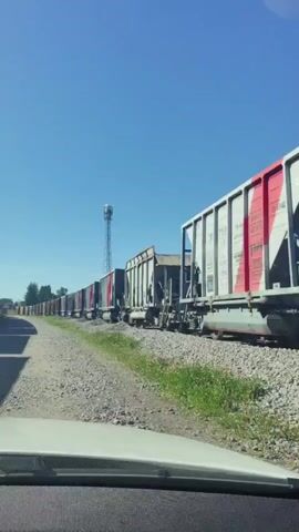 The video shows a long train with many open-top hopper cars moving along a track. The train is seen from inside a vehicle, suggesting it is moving parallel to the train. The environment appears to be rural or industrial, with a clear blue sky and some greenery alongside the tracks. A tall communications tower is visible in the distance. The train cars are mostly gray, with some having red and white markings. Some cars appear empty, while others may contain material. The camera perspective is low, focused on the dashboard of the vehicle.