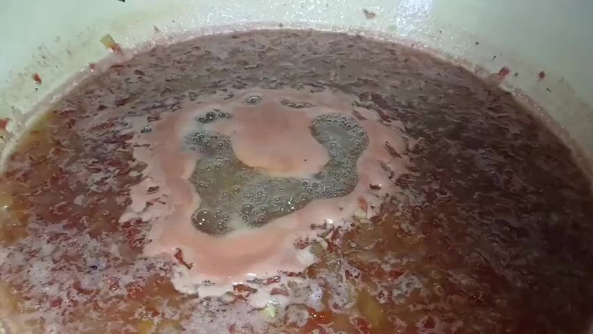 The video shows a close-up, overhead view of a pot of boiling liquid. The liquid is a reddish-pink color and appears to be chunky, possibly with pieces of fruit or vegetables. A layer of foam is present on the surface, with a larger, circular area of bubbling and foam in the center. The pot itself is a light color, possibly white or cream, and its rim is visible at the top and sides of the frame. The lighting is even and appears to be from overhead. No people or other objects are visible in the frame. The focus is entirely on the contents of the pot.
