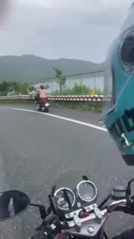 The video is shot from the perspective of a person riding a motorcycle on a road with mountains in the background. The road appears to be wet, and the sky is overcast, suggesting it might be raining or has recently rained. The motorcycle has a red gas tank and a small windscreen. The rider is wearing a turquoise helmet with a white logo. In the first few seconds, another motorcycle with two people on it is visible on the opposite side of the road. The motorcycle rider then overtakes it, and it is seen again a short distance ahead. The scenery consists of green vegetation along the roadside and the mountains in the distance.