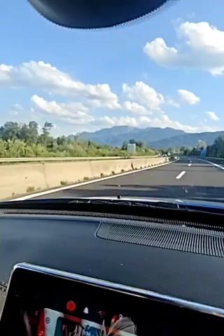 The video shows a first-person perspective of driving on a highway on a clear day. The road is paved with white lines marking the lanes. There is a concrete barrier separating the lanes. On the left side of the road, there are trees and in the distance, mountains under a blue sky with scattered clouds. A white van is visible driving in the opposite direction. The dashboard of the car is partially visible at the bottom of the frame, with a black surface and a speaker grille.