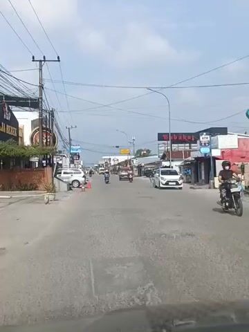 The video shows a street scene with several cars, motorcycles, and buildings on either side of the road. The sky is cloudy, and there are many power lines overhead. On the left side of the road, there are several businesses, including one with a sign that says "FOOMEE." There are also some cones on the side of the road. On the right side of the road, there are more businesses, including one with a sign that says "BABAKAR." There is also an ATM machine visible. The traffic appears to be light, with only a few vehicles moving at a time. The overall impression is that of a typical street in a commercial area.