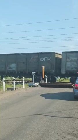 A train made of gray freight cars is passing over a railroad crossing. The crossing has a red and white striped barrier and black and white striped posts. The train cars have Cyrillic writing on them. The sky is clear and blue, and there are overhead wires for the train. The camera is positioned inside a vehicle, as evidenced by the windshield and rearview mirror. The video is shot from a slightly low angle.