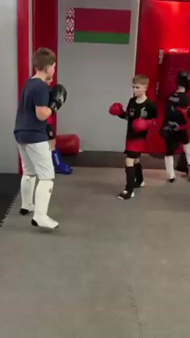 In a gym, two young boys are sparring in what appears to be a kickboxing or martial arts session. The boy on the left wears a navy T-shirt, light gray shorts, and white shin guards, with black boxing gloves. His opponent wears a black and red uniform with matching red gloves and black shin guards. They exchange punches under the watchful eye of an adult, with the Belarusian flag displayed prominently in the background.