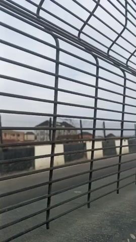 The video shows a close-up of a metal fence with a blurred view of a road and buildings in the background. The road has black and white striped barriers along the side. Traffic noise can be heard. The sky is overcast.