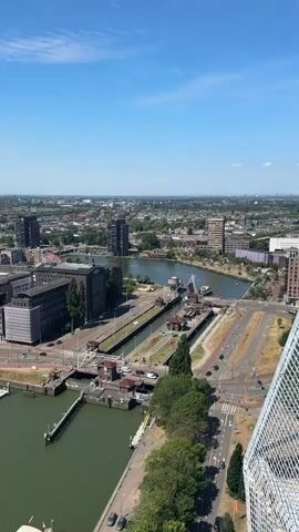 This is a high-angle aerial shot of a city on a clear, sunny day, likely midday given the bright sunlight. The scene is a waterfront with a lock system in the foreground, connecting a larger body of water to a canal or smaller waterway. Several boats are docked in a marina to the left.

The city features a mix of modern and older architecture, with several tall, dark-colored apartment buildings and wider, lower-rise commercial or residential structures. Roads with cars are visible, indicating an active urban environment. The landscape extends into the distance, with more buildings and greenery under a bright blue sky with scattered white clouds. The overall impression is one of a well-developed, bustling European city.

There are no visible people in the frame. The focus is on the urban landscape, the waterways, and the lock system. The weather appears to be warm and pleasant, contributing to a vibrant and clear view of the city. The presence of the lock system suggests a port or a city with significant water transportation infrastructure.