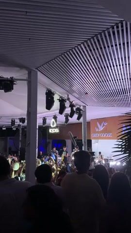 The video shows a crowded indoor venue, likely a nightclub or concert hall, bathed in red and purple lighting. The ceiling is lined with horizontal white slats, and professional stage lighting hangs from the structure. A band is performing on stage, with several members visible playing instruments. Prominent logos for "IQOS" and "Grey Goose" are displayed on screens behind the stage. The audience is densely packed, with many people holding up their phones, presumably to record the event. The overall atmosphere appears to be energetic and celebratory.