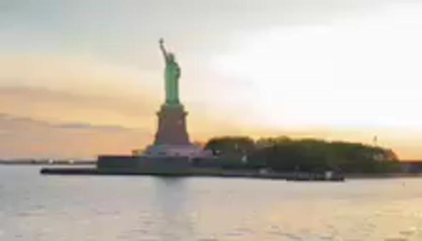 This media file shows the Statue of Liberty on Liberty Island in New York Harbor. The statue is green, indicating its copper exterior has oxidized. It stands on a pedestal on a man-made island, with trees visible behind it. The statue is silhouetted against a bright, hazy sky, suggesting it is either sunrise or sunset. The sun appears to be setting, casting a warm, orange glow on the clouds. The water in the foreground is calm, reflecting the sky. The overall scene evokes a sense of peace and grandeur. No people are clearly visible, though there might be some indistinct figures on the island. The location is identifiable as Liberty Island, New York, due to the iconic statue. The time of day is clearly either dawn or dusk due to the lighting conditions and the colors in the sky. The weather appears clear and calm.