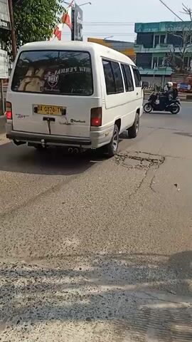 A white van is driving on a road with potholes. The van has a license plate that reads "DA 0876 TH". A person on a motorcycle is riding behind the van. There are buildings and trees in the background. The sky is clear.