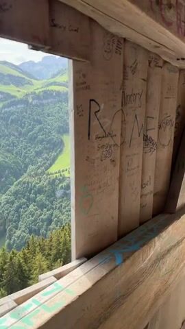 The video begins inside a small wooden structure with graffiti-covered walls. The camera then pans to reveal an outdoor wooden walkway clinging to the side of a steep cliff. A metal railing runs along the walkway's edge. In the distance, there are lush green hills and a large mountain with patches of snow on its peak. The sky is bright with some clouds. A person can be seen walking in the distance on the walkway.