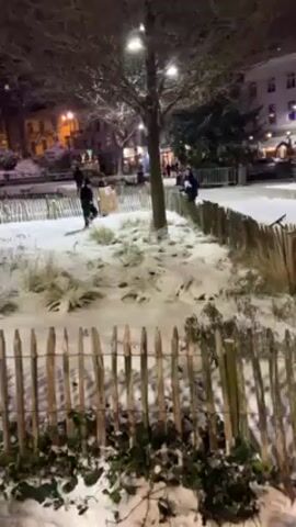 Two children are having a snowball fight in a snow-covered town square at night. The square is surrounded by a wooden fence and trees, and there are buildings in the background lit with streetlights. One child, wearing a black coat and a hat, throws a snowball at the other child, who is standing behind the fence. The camera is positioned behind the fence, looking into the square. The scene is lit by streetlights, creating a cold, wintery atmosphere.