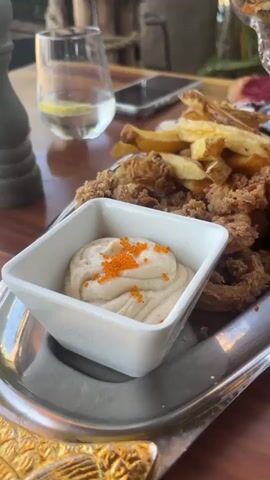 A close-up, slightly shaky, eye-level video pans across a platter of fried seafood and accompaniments. In the foreground, a white ceramic square dish contains a creamy dip, possibly aioli or mayonnaise, garnished with bright orange roe. Adjacent to this dish, a large portion of fried calamari rings and pieces sits piled high. Behind the calamari, a heap of golden french fries is visible. Further back on the platter, several mussels in their shells, coated in a creamy sauce, are arranged. To the right of the mussels, a couple of large prawns, their shells on, are displayed. To the right of the prawns, several pieces of flatbread, some with visible orange roe, rest on a red plate. In a tall, stemmed glass bowl at the back of the platter, small fried fish, also garnished with orange roe, are presented. A slice of lemon rests on the platter near the mussels. In the background, a glass with water and a lemon slice, a smartphone, and a blurry outdoor environment with trees and gravel are visible. The platter itself is silver and rests on a wooden table.