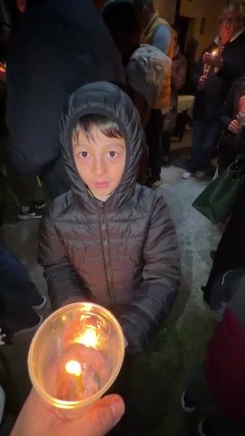 A young boy in a black hooded puffer jacket is holding a lit candle in a plastic cup. The camera focuses on the candle, then shifts to the boy's face, and then zooms in on the flame being transferred to his hands. The camera then focuses on the boy's jacket. The scene appears to be outdoors and it is dark, with other people holding candles in the background.