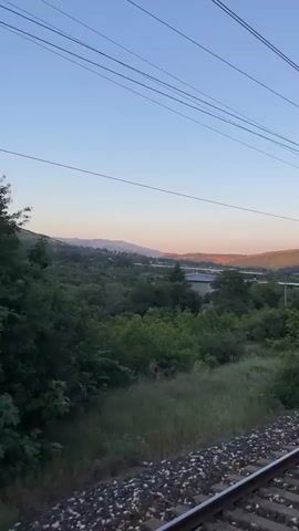A view from a train as it travels through a rural landscape. The sky is clear and blue with a hint of orange from the setting sun. There are trees and hills in the background. Overhead power lines for the train are visible. The train tracks are in the foreground. The video is shot from the perspective of someone looking out a window on the right side of the train.