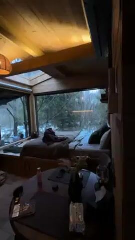 A shirtless man leaps from the side of a daybed platform, landing awkwardly on the floor in a cozy wooden room with a large picture window overlooking a snowy landscape. He moans and lies on his back. The room features wood flooring and paneling, a round table with assorted items, a pendant lamp, a fireplace with stacked wood, and a door leading to an outdoor area.