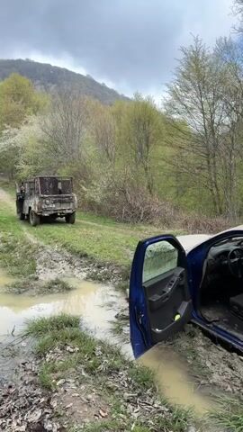 The video captures a rural, natural scene on a cloudy day, likely in the Manavi, Georgia region. In the foreground, a blue SUV, which appears to be a Nissan Xterra, is stuck in a deep, muddy puddle on a dirt track. The driver's side door of the SUV is wide open, revealing part of its interior and emphasizing its predicament in the mud.

Further up the muddy track, an older, rugged-looking utility truck, possibly military-style with a green or camouflage paint job and a high-sided cargo bed, is parked or waiting. A person is visible in the driver's seat of this truck. The truck appears to be on a slightly higher, less submerged part of the track.

The surrounding landscape is characterized by dense vegetation, including trees with fresh green leaves, indicating springtime. Rolling hills or mountains are visible in the background under an overcast sky. The ground is visibly wet and muddy, with significant puddles and ruts, suggesting recent rainfall or generally damp conditions, common for off-road trails. The overall scene depicts an off-road challenge, with one vehicle stuck and another nearby, possibly in a convoy or assisting.