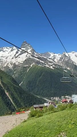 The video shows a scenic mountain view from a ski lift. The ski lift chairs are visible as they ascend the steep, green, grassy slope. The background features snow-capped mountains, lush green forests, and a clear blue sky. In the foreground, a person wearing a white jacket, a pink baseball cap, and sunglasses is seen resting on the ski lift seat. The person is looking up and smiling, enjoying the view. The ski lift appears to be operational and carrying passengers up the mountain.