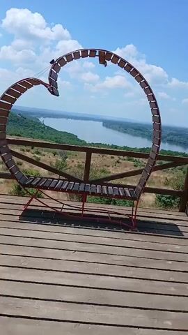 A heart-shaped bench with wooden slats and a red metal frame is positioned on a wooden deck. The bench overlooks a wide river surrounded by rolling green hills and a blue sky with white clouds. The camera pans upwards to reveal the expansive view.