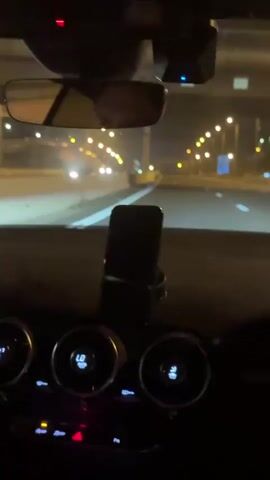The video begins with a blurry shot from inside a car at night, looking out the windshield. The interior of the car, including the dashboard with glowing circular displays and buttons, is dark. Outside, a highway is visible with streetlights lining the road, creating a bokeh effect. As the video progresses, the view outside becomes slightly clearer, showing the car in motion. The streetlights continue to illuminate the highway, with other vehicles visible in the distance. The camera remains focused on the forward view, capturing the experience of driving at night.