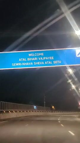 The video shows a nighttime view from a vehicle traveling on a highway. The vehicle passes under a blue overhead sign that reads "WELCOME ATAL BIHARI VAJPAYEE SEWRI-NHAVA SHEVA ATAL SETU". The highway is well-lit by streetlights on tall poles. Barriers separate the lanes, and there are white lane markings on the asphalt road. The sky is dark, indicating it is nighttime, and there are some visible light reflections on the windshield or camera lens.