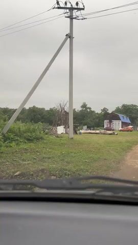 A video shot from inside a vehicle shows a rural scene under a cloudy sky. In the foreground, the top of a car's dashboard and windshield wipers are visible. The middle ground features a grassy area with a large pile of branches in the shape of a teepee, some scattered wooden planks, and a large log. Further back, a red and white barn with a blue roof stands with an orange vehicle parked nearby. Trees form a dense line in the background. A utility pole with wires is prominent to the left of the frame. The camera is stationary throughout the short clip.