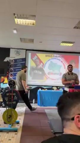 The video shows a room set up for an arm wrestling competition. In the foreground is a red arm wrestling table, with a referee standing behind it, wearing a striped shirt and dark pants. He is gesturing with his hands and appears to be addressing the crowd.

To the left of the referee, a man in a green shirt is walking toward the table. To the referee's right, a man with a beard, wearing a white tank top and dark pants, stands listening to the referee. Behind them, several other people are standing and watching.

Behind the referee, a large projection screen displays the competition's logo, which reads "BRAS GAUCHE, FEDERATION FRANCAISE DE FORCE." The room is well-lit with fluorescent ceiling lights. To the left of the table, there is weightlifting equipment. In the foreground is a man in glasses with a blurred focus.