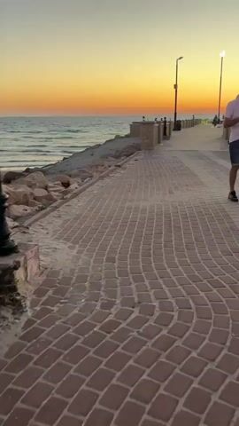 A person walks along a brick pathway at sunset by the ocean, extending their hand towards the camera, showing off their painted nails, before continuing to walk along the pathway, past a pier with two statues of people dancing and streetlights.