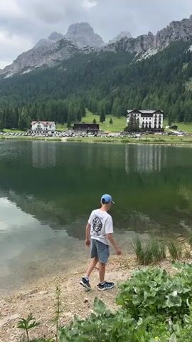 A young boy wearing a white t-shirt, blue cap, and denim shorts stands at the edge of a clear lake, looking out at the water. The lake reflects a lush green forest and imposing mountains under a cloudy sky. In the background, several buildings, possibly hotels, are situated near the tree line, with cars parked in front of them. The foreground shows the rocky shoreline of the lake with some green vegetation. The overall scene is a scenic mountain landscape with a boy enjoying the view.